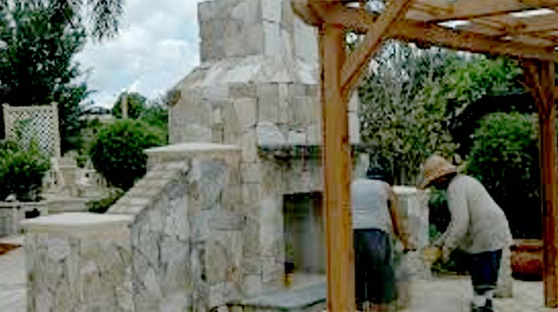 <Here are Home Services For You experts constructing an outdoor stone fireplace in a backyard setting. The structure is made from natural, irregularly shaped stone pieces, giving it a rustic appearance.  The fireplace is integrated into a landscaped area.>